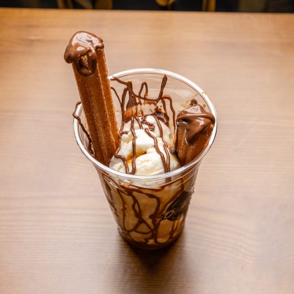 A clear cup of vanilla ice cream with chocolate syrup and two churros on a wooden table.