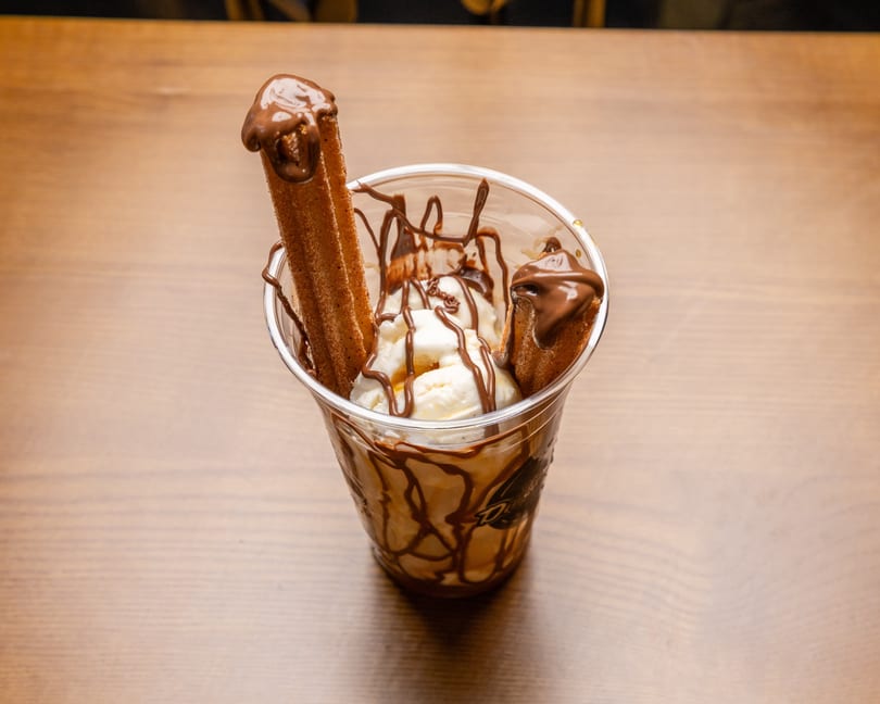 A clear cup of vanilla ice cream with chocolate syrup and two churros on a wooden table.