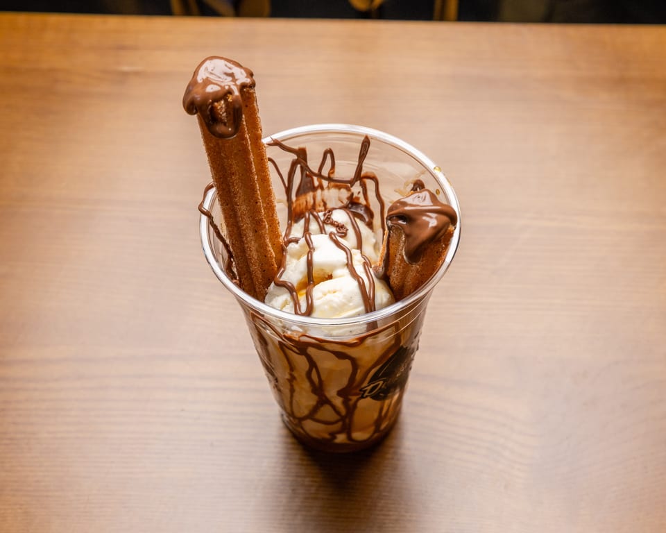 Vanilla ice cream with chocolate drizzle and two churros in a clear plastic cup.